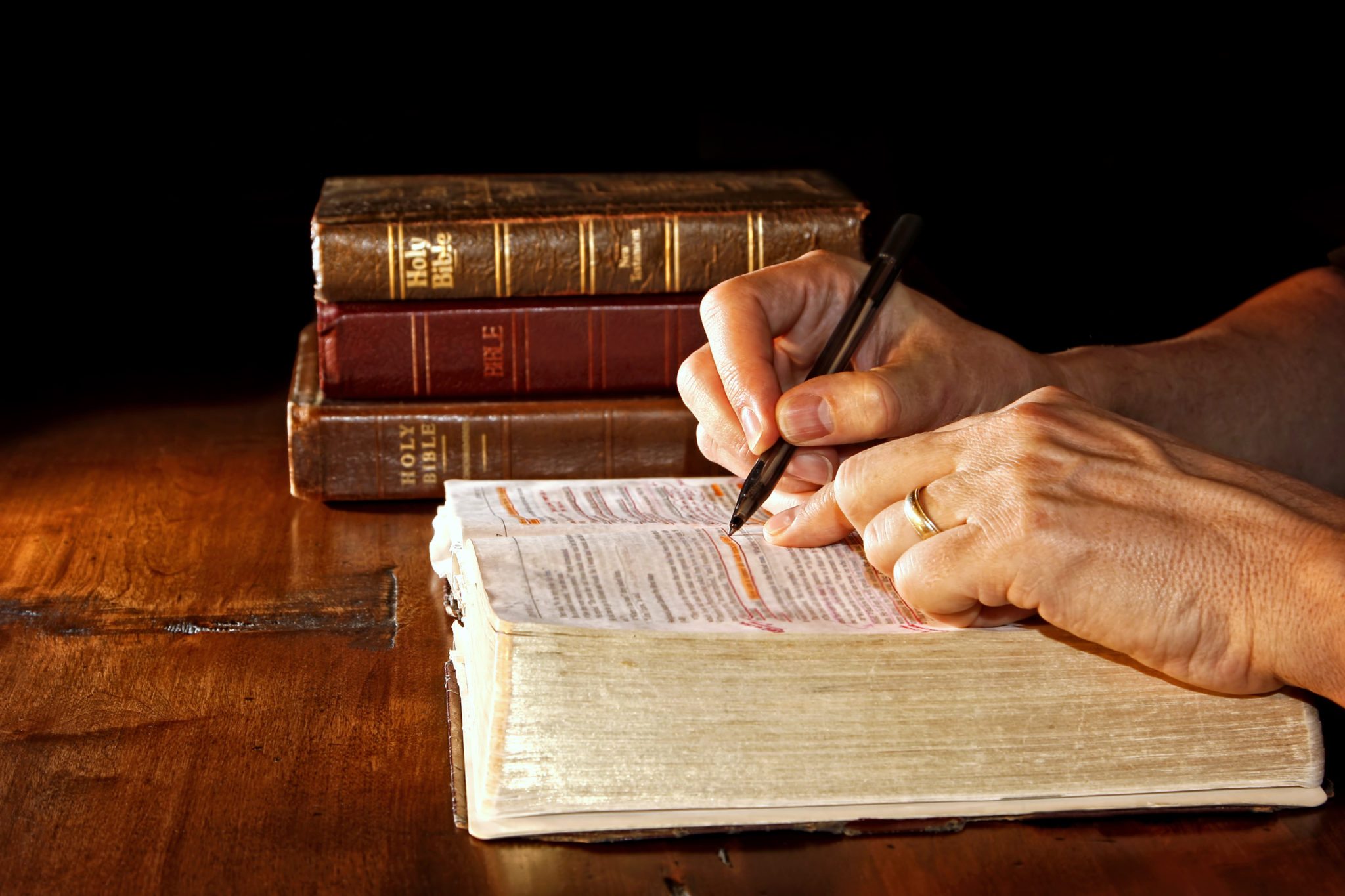 A man uses a pen to help him study an old and worn Holy Bible while other versions and/or translations of the bible are nearby on the wood table.